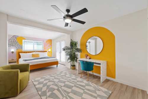 Brightly decorated bedroom featuring a large bed, colorful wall art, a desk with a mirror, and a cozy chair.