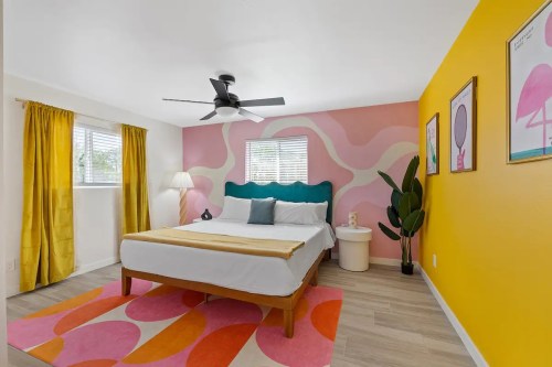 Brightly colored bedroom with a large bed, yellow curtains, a fan, and playful wall art in pink and orange tones.