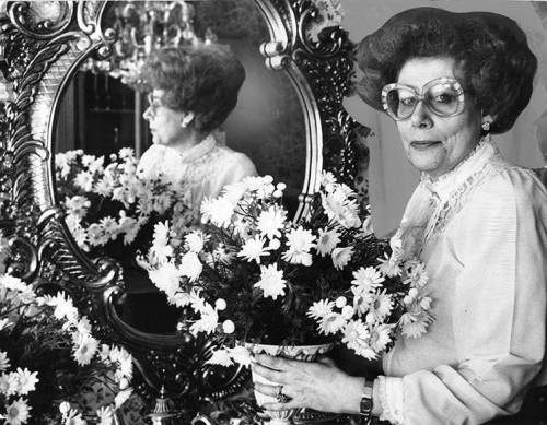 A woman with large glasses holds a vase of flowers, reflected in an ornate mirror. Black and white photo.