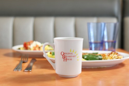 A white coffee mug with "Original Breakfast House" logo in front of a plate of breakfast food and two glasses of water.