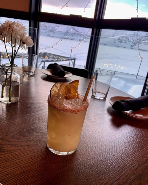 A refreshing cocktail with a dried orange slice sits on a wooden table, overlooking a snowy landscape through large windows.