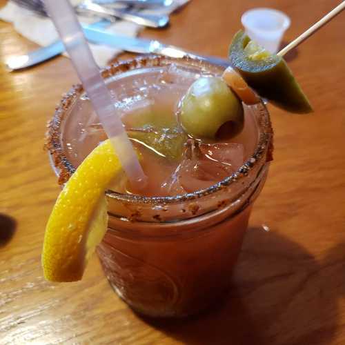 A close-up of a cocktail in a glass, garnished with a lemon slice, olives, and jalapeños, on a wooden table.