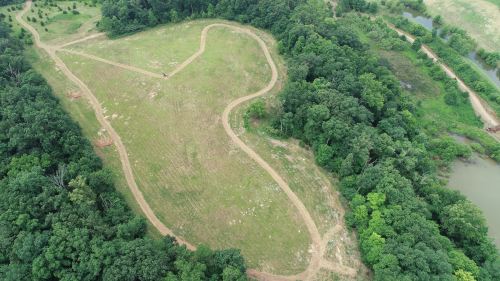 Aerial view of a winding dirt path through a green landscape with trees and a small body of water nearby.