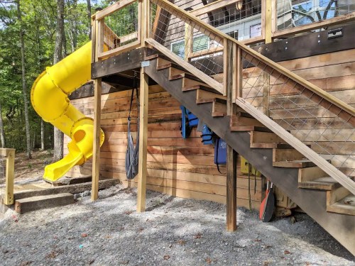 A wooden structure with a yellow slide and stairs, surrounded by trees and gravel.