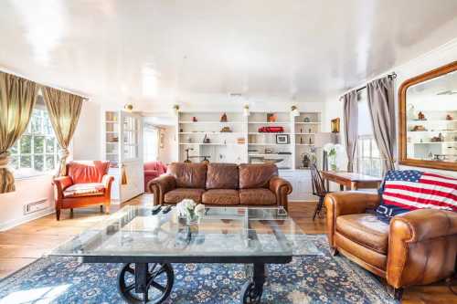 Cozy living room with leather sofas, a glass coffee table, and shelves displaying decor, illuminated by natural light.