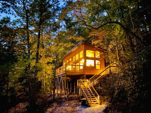 A modern wooden cabin elevated on stilts, surrounded by trees, illuminated warmly at dusk.