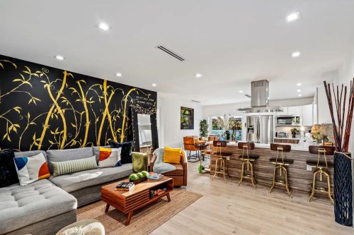 Modern living room with a gray sectional, bamboo wall art, and a kitchen bar with stools. Bright and stylish decor.