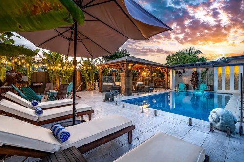 A serene pool area with lounge chairs, an umbrella, and vibrant sunset skies, surrounded by lush greenery and cozy seating.