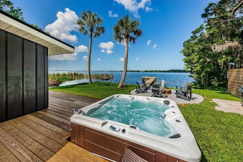 A hot tub on a wooden deck overlooking a lake, surrounded by palm trees and outdoor seating. Bright blue sky above.