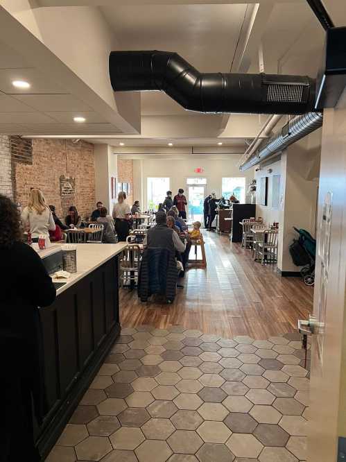 A bustling restaurant interior with patrons dining, featuring wooden floors and a mix of tables and chairs.