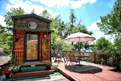 A cozy tiny house with a wooden deck, surrounded by greenery, featuring a table, chairs, and a large umbrella.