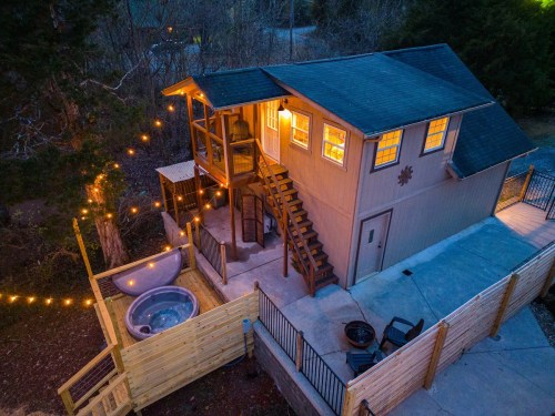 A cozy cabin with a hot tub, string lights, and a staircase, surrounded by trees in a serene outdoor setting.
