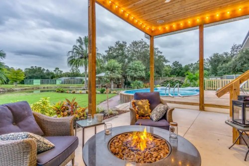 Cozy outdoor patio with a fire pit, seating, and a pool surrounded by lush greenery under a cloudy sky.