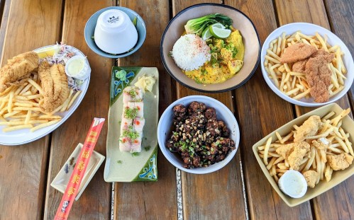 A variety of dishes on a wooden table, including fried fish, rice, noodles, and fries, with sauces on the side.