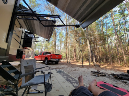 A view from an RV campsite with a red truck, trees, and a person relaxing in a chair with bare feet visible.