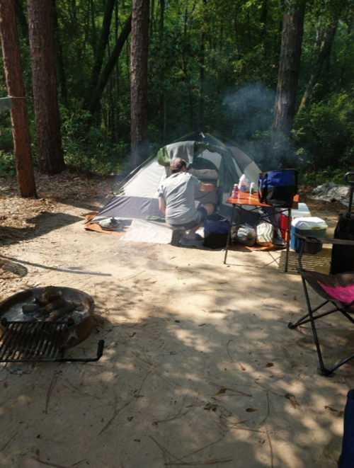 A person prepares food outside a tent in a wooded campsite, with smoke rising from a nearby fire pit.