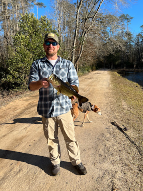 A man in a plaid shirt holds a large fish on a dirt path, with a dog standing nearby and trees in the background.