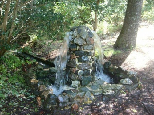 A stone fountain surrounded by greenery, with water flowing from the top into a circular basin.
