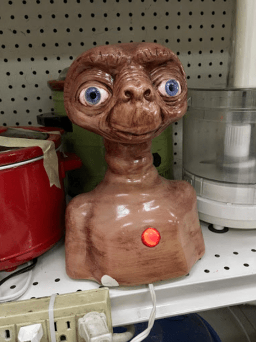 A quirky E.T. figurine with large eyes and a glowing red button, displayed on a shelf among kitchen appliances.