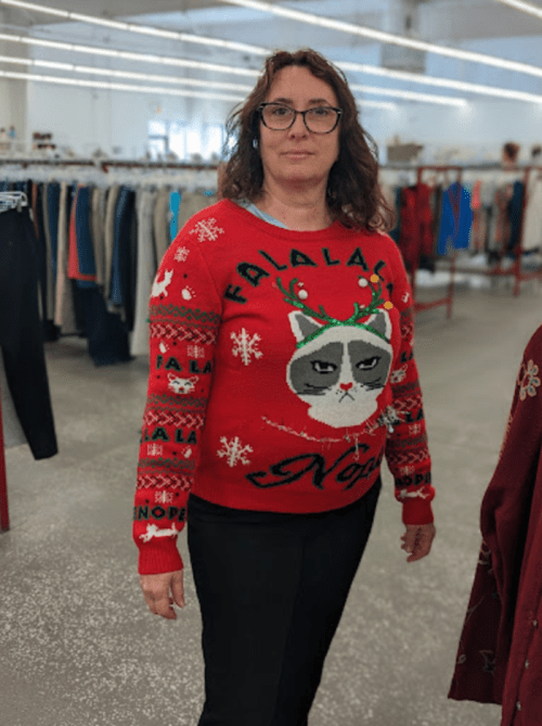 A woman wearing a festive red cat-themed sweater stands in a clothing store aisle filled with clothes.