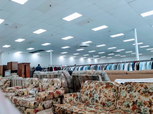 A spacious thrift store interior with rows of clothing racks and floral-patterned sofas in the foreground.