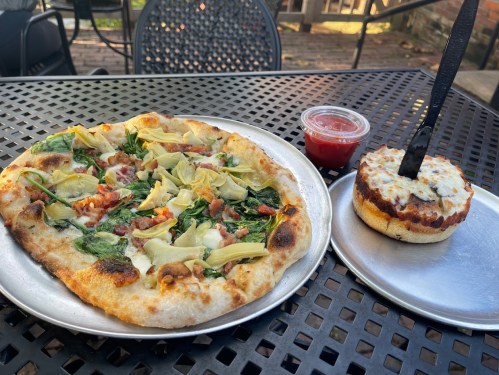 A pizza topped with spinach, artichokes, and bacon, alongside a small dish of marinara sauce and a cheesy bread.