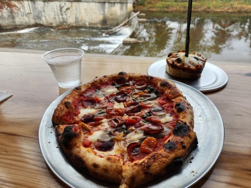 A delicious pepperoni pizza on a metal plate, with a glass of water and a small dessert, by a scenic water view.