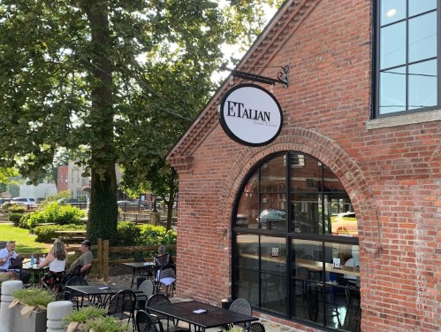 Exterior of a brick restaurant with a sign reading "ETALIAN," featuring outdoor seating and trees in the background.