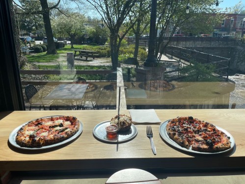 Two pizzas on a table by a window, with a view of a park and a stream outside. Sunlight brightens the scene.