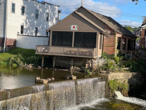A riverside building with a porch overlooks a small waterfall, surrounded by greenery and a clear blue sky.