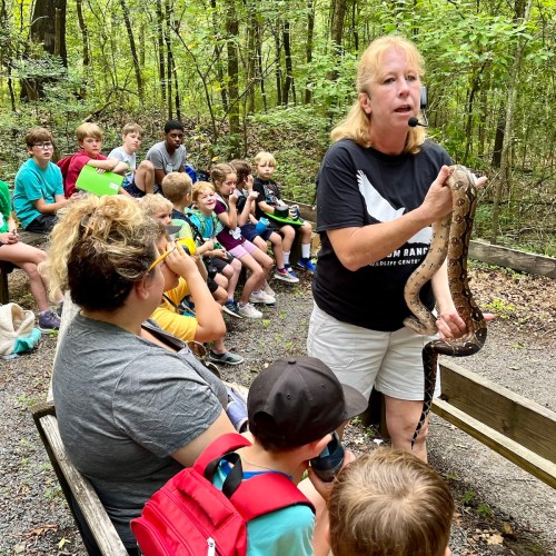A woman holds a snake while speaking to a group of children in a forested area.