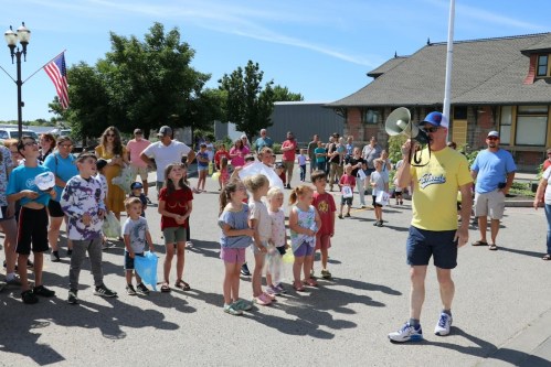 A crowd of children and adults gathers outdoors as a man speaks into a megaphone, with flags and buildings in the background.