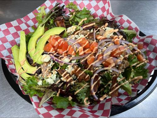 A colorful salad with greens, avocado, tomatoes, red onions, bacon, and drizzled sauce in a checkered basket.