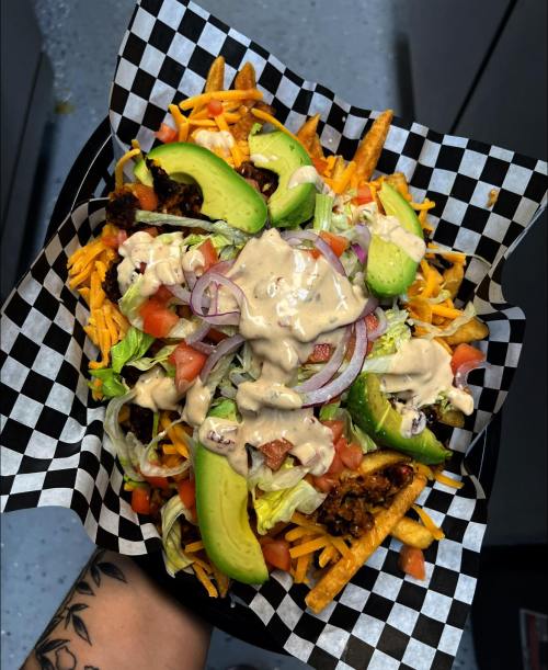 A hand holding a checkered basket filled with loaded fries, topped with lettuce, tomatoes, onions, avocado, and sauce.