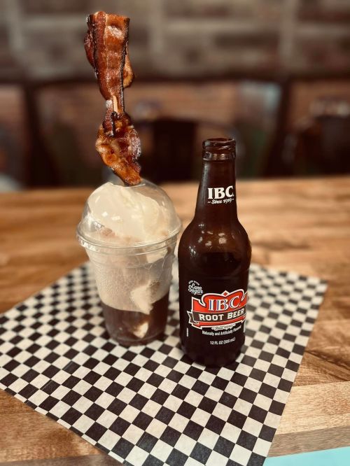 A root beer bottle next to a dessert cup topped with ice cream and a piece of crispy bacon.