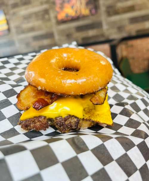 A burger with a glazed donut as the bun, featuring cheese and bacon, served on a checkered paper.