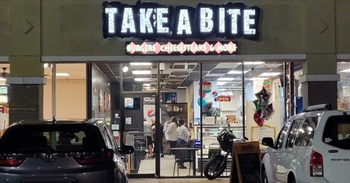 Exterior of "Take A Bite" restaurant, featuring a sign and customers inside, with cars parked outside.