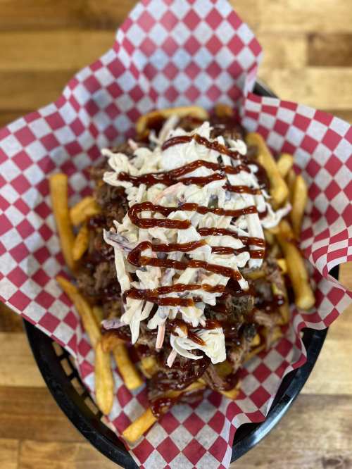A basket of fries topped with pulled pork, coleslaw, and barbecue sauce, served in a red and white checkered paper.