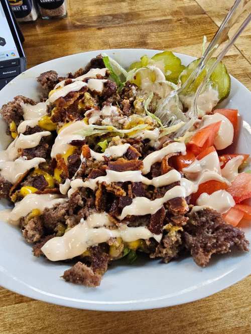 A bowl of salad topped with ground beef, cheese, bacon, tomatoes, pickles, and drizzled with a creamy dressing.
