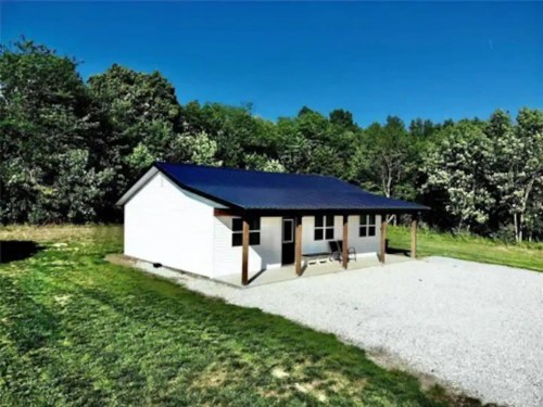 A small white house with a blue roof, set on a gravel driveway surrounded by green trees and clear blue sky.