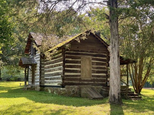 A rustic log cabin surrounded by trees and grass, featuring a boarded window and a sloped roof.