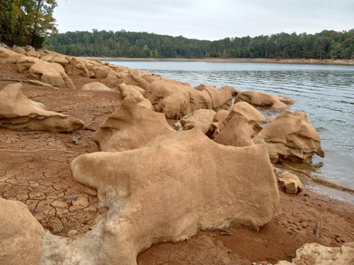 Rocky shoreline with unique, weathered formations along a calm lake, surrounded by trees and a cloudy sky.