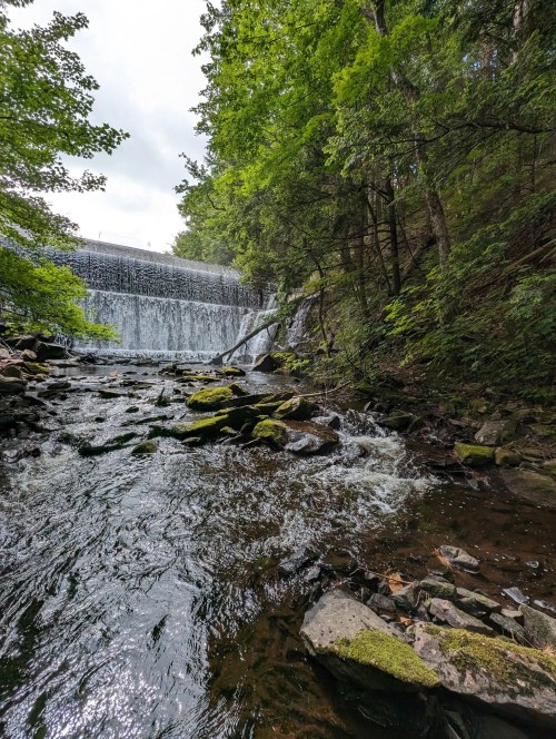 A serene waterfall cascades over rocks, surrounded by lush green trees and a calm stream below.