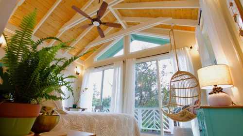 Cozy bedroom with a hanging chair, large windows, and wooden ceiling, surrounded by greenery and soft natural light.