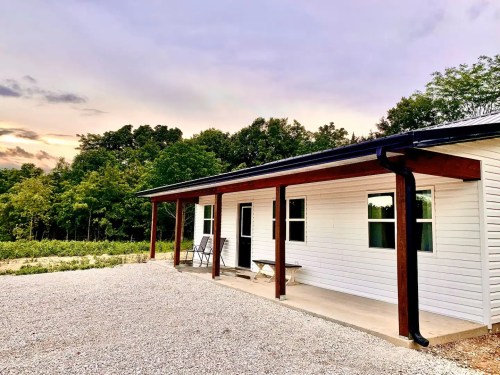 A modern white house with a porch, surrounded by trees and a gravel pathway, under a colorful sunset sky.