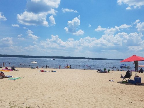 A sandy beach with people swimming and relaxing under a blue sky filled with clouds, and a red umbrella in the foreground.