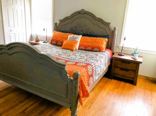 A cozy bedroom featuring a gray bed with an orange patterned blanket, two nightstands, and potted plants.