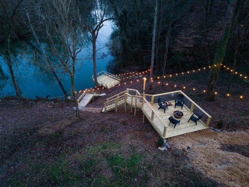 A serene riverside area with wooden decks, chairs, and string lights, surrounded by trees and water.