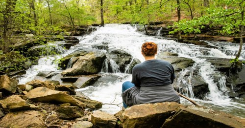 A person sits on rocks by a flowing waterfall, surrounded by lush greenery in a serene forest setting.