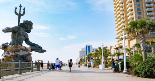A coastal boardwalk with a large statue of Neptune, palm trees, and people walking along the path.